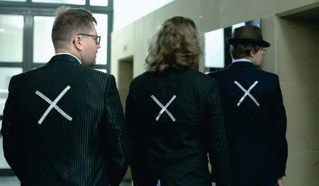 The image shows three individuals walking away from the camera in a corridor. They are dressed in formal attire, with the two on the left wearing black suits and the one on the right wearing a dark suit with a hat. Each person has a large white "X" drawn on the back of their jacket. The corridor has a modern design with glass doors and a neutral color scheme. The individuals appear to be in a professional setting, possibly an office or a corporate building. The lighting is bright, and the overall atmosphere is serious and somewhat mysterious due to the "X" markings.
