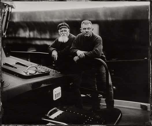 Plymouth RNLI Coxswains Dave Milford and Sean Marshall leaning side-by-side on a rail on the foredeck of the station’s Severn class all-weather lifeboat. The superstructure of the 42 tonne, 17 metre vessel encroaches from the edge of the frame on the left hand side and the dock wall is out of focus behind them. Dave is in his late sixties and is wearing a traditional RNLI cap. He has a long white beard. Sean is in his fifties and they’re both wearing traditional RNLI Ganseys. The black and white photograph was handmade on 12x10 inch glass by Jack Lowe using a process called wet collodion (or wet plate), which was invented in 1851. 