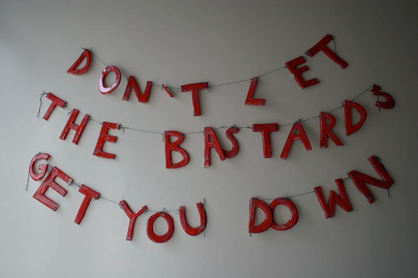 A photograph captures a three-tiered banner made of large, hand-painted red metal letters strung on thin wires against a plain light-grey wall. The letters are arranged in gentle upward arcs, reading "DON'T LET" on the top row, "THE BASTARDS" on the middle row, and "GET YOU DOWN" on the bottom row. The letters have a slightly distressed, artisanal appearance.