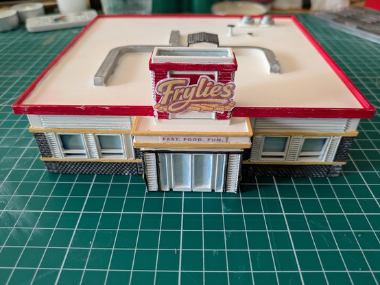 A plastic model of the Frylies fast food restaurant sits on a green, white-lined cutting mat. The top most part of the building is ringed in red with large signage out front. Over the door are the words "Fast. Food. Fun."