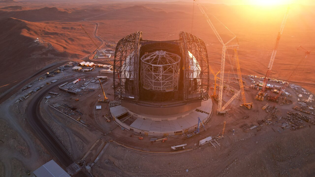 A drone photograph of a gigantic telescope in the desert, at sunset. The telescope dome is under construction, and partially covered in cladding. The sliding doors of the dome are open, revelaning the telescope inside: a cylindrical structure of white criss-crossed beams. The dome is surrounded by huge cranes, and the entire scene is bathed in the golden light of the setting Sun in the background.