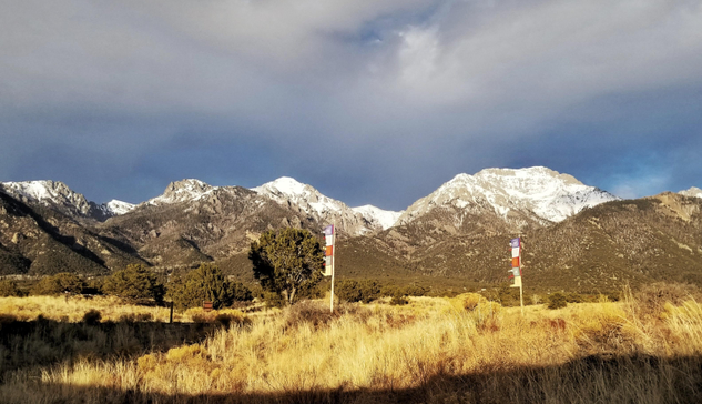 Sunset light casts a golden hue on dried grasses, brush, and Piñon Pines, while lighting up a snow-covered mountain range. There are two vertical Tibetan prayer flags on wooden poles blowing in the wind, colored the traditional blue, white, red, green, and yellow colors.