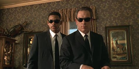 Still from the movie _Men In Black_, during their interview with 'Beatrice' (whose husband has just been taken over by a bug). Shown: Will Smith (left, slightly behind) and Tommy Lee Jones (right, slightly forward) standing in a dark, formal living room. Both are wearing their trademark dark glasses.