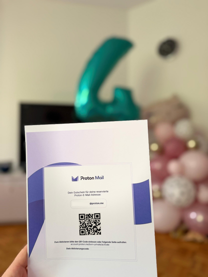 A person’s hand holding a Proton Mail ‘Born Private’ gift certificate. The card shows the Proton Mail logo, a reserved @proton.me email address, a large QR code and activation instructions. In the background there are colorful party balloons, suggesting a baby celebration.