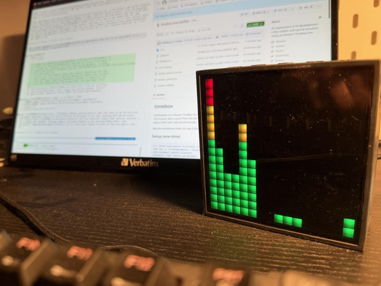 A screen in the background with almost visible coding agent and the github page of the project. The Divoom Timebox Evo is in front showing the green, yellow and red pixels of the sound visualizer.